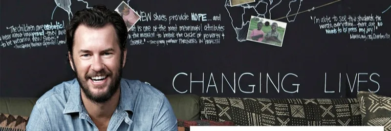 Ο Blake Mycoskie, ιδρυτής της TOMS, χαμογελά μπροστά από έναν μαυροπίνακα.