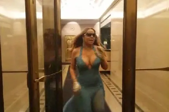 Η Mariah Carey με γαλάζιο φόρεμα και γυαλιά ηλίου σε διάδρομο κτιρίου.