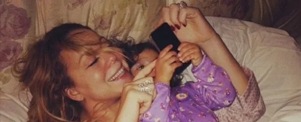 Η Mariah Carey ξαπλωμένη στο κρεβάτι με την κόρη της, κρατώντας ένα κινητό τηλέφωνο.