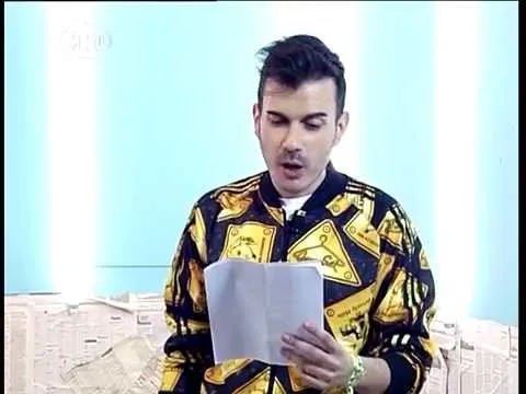 Παρουσιαστής με μοτίβο ζακέτα διαβάζει χαρτί σε τηλεοπτικό στούντιο.