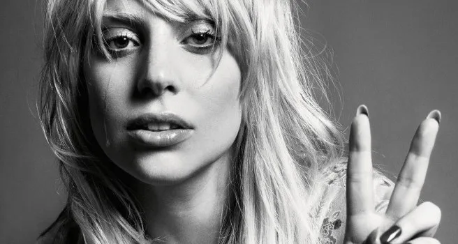 Ασπρόμαυρο κοντινό πορτρέτο της Lady Gaga με ξανθά μαλλιά και αφέλειες, κάνοντας το σήμα της ειρήνης.
