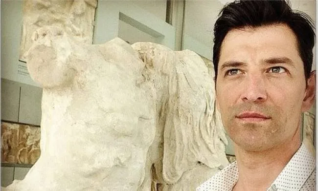 Ο Σάκης Ρουβάς ποζάρει δίπλα σε αρχαίο γλυπτό σε μουσείο.