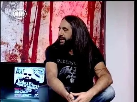Παρουσιαστής με μακριά μαλλιά και μαύρη μπλούζα σε τηλεοπτικό πλατό.