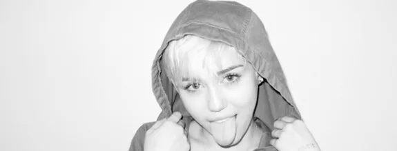 Η Miley Cyrus με κουκούλα βγάζει τη γλώσσα της έξω σε ασπρόμαυρη φωτογραφία.