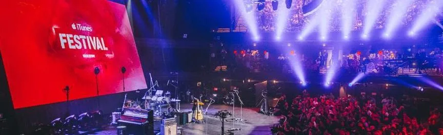 Σκηνή του iTunes Festival με μεγάλη κόκκινη οθόνη και μουσικά όργανα.