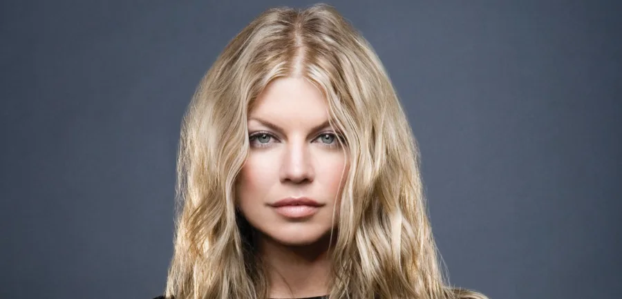 Κοντινό πορτρέτο της Fergie με ξανθά κυματιστά μαλλιά σε γκρι φόντο.