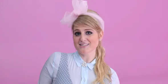 Η Meghan Trainor με ροζ κορδέλα στα μαλλιά και γαλάζιο πουκάμισο σε ροζ φόντο.