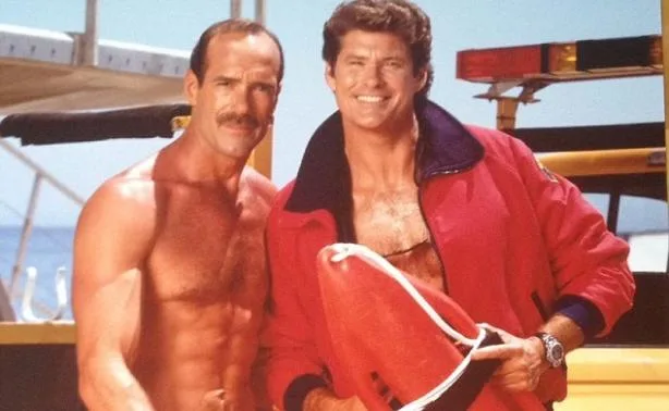 Ο David Hasselhoff με κόκκινο μπουφάν ναυαγοσώστη δίπλα σε έναν άνδρα χωρίς μπλούζα.