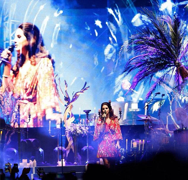 Η Lana Del Rey τραγουδάει ζωντανά σε σκηνή με τροπικά δέντρα και κεριά.