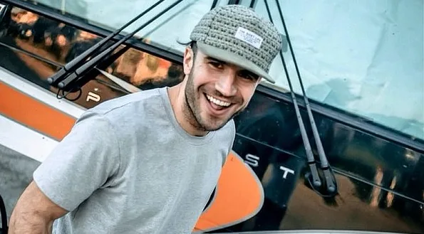 Ο Sam Hunt χαμογελάει φορώντας γκρι μπλουζάκι και καπέλο μπροστά από ένα λεωφορείο.