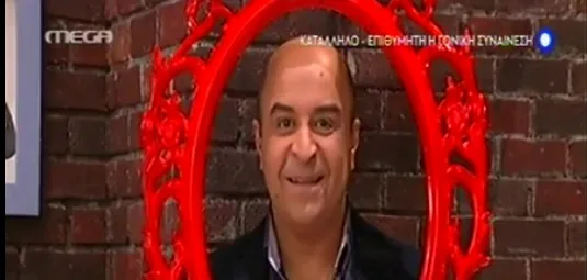 Ο Μάρκος Σεφερλής κρατάει μια κόκκινη κορνίζα μπροστά από το πρόσωπό του.