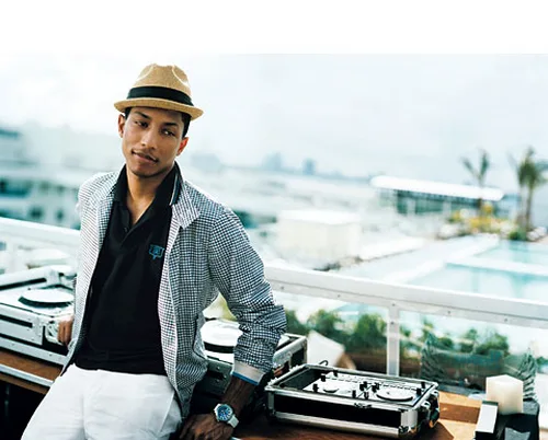 Ο Pharrell Williams με καπέλο και καρό τζάκετ μπροστά από εξοπλισμό DJ.