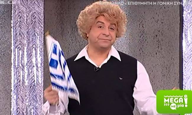 Ο Μάρκος Σεφερλής με ξανθιά περούκα κρατώντας την ελληνική σημαία.