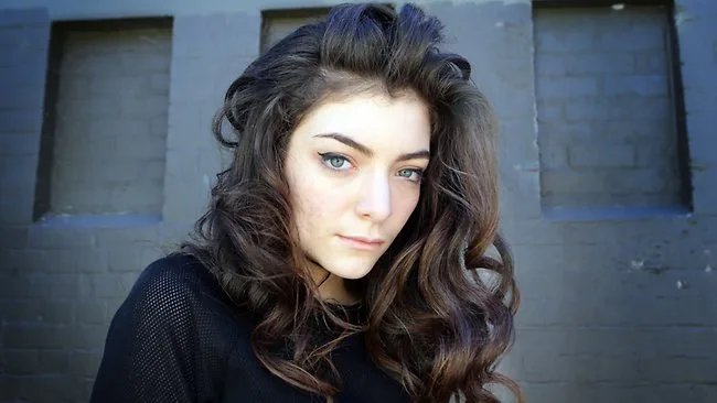 Η Lorde με μακριά σγουρά μαλλιά μπροστά από έναν γκρίζο τοίχο.