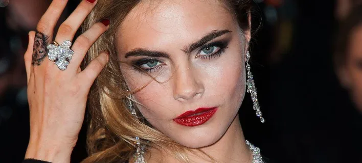 Κοντινό πλάνο της Cara Delevingne με έντονο μακιγιάζ και κοσμήματα.