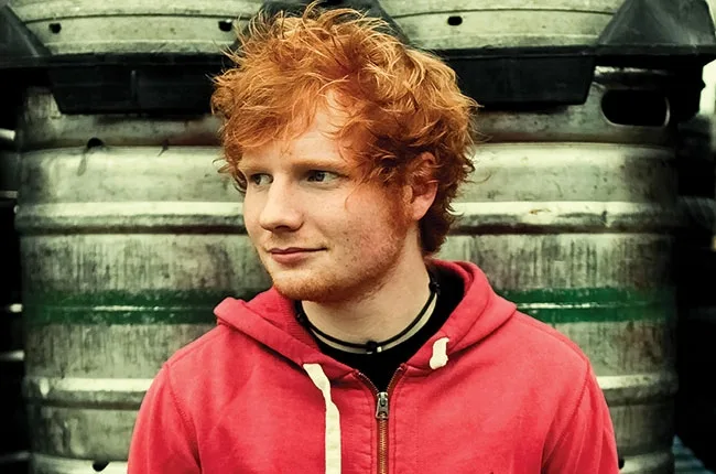 Ο Ed Sheeran με κόκκινο φούτερ μπροστά από μεταλλικά βαρέλια.
