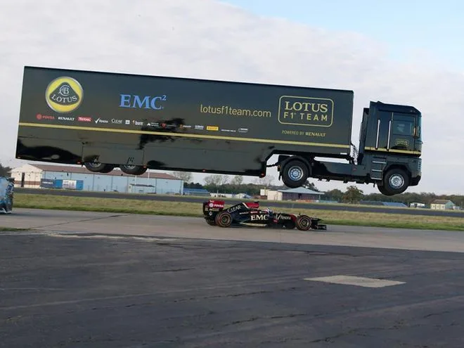 Ένα μεγάλο μαύρο φορτηγό της Lotus F1 Team πραγματοποιεί άλμα πάνω από ένα μονοθέσιο της Formula 1.