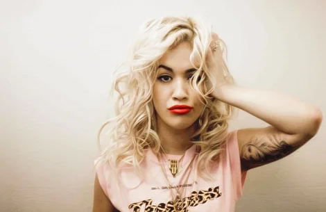 Η Rita Ora με ξανθά σγουρά μαλλιά και κόκκινο κραγιόν.