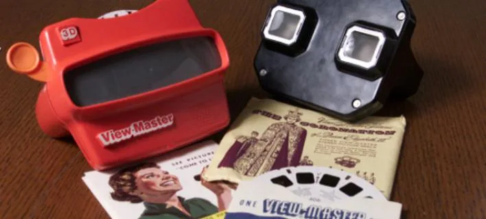 Δύο συσκευές View-Master, μία κόκκινη και μία μαύρη, μαζί με δίσκους εικόνων και συσκευασίες πάνω σε ξύλινη επιφάνεια.