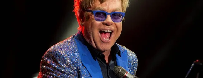 Ο Sir Elton John με μπλε γυαλιά ηλίου και αστραφτερό σακάκι.