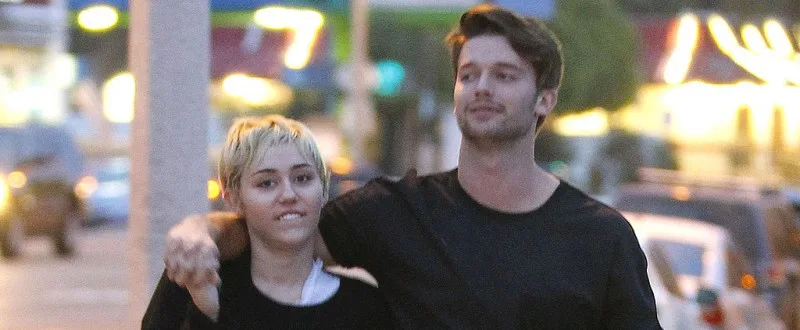 Η Miley Cyrus και ο Patrick Schwarzenegger περπατούν αγκαλιασμένοι σε εξωτερικό χώρο.
