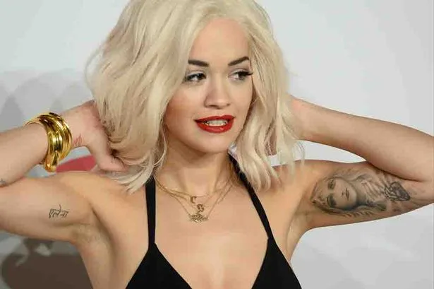 Η Rita Ora με ξανθά μαλλιά και κόκκινο κραγιόν ποζάρει με τα χέρια πίσω από το κεφάλι.