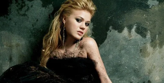 Η Kelly Clarkson ποζάρει με μαύρο φόρεμα σε φόντο με φθαρμένο τοίχο.