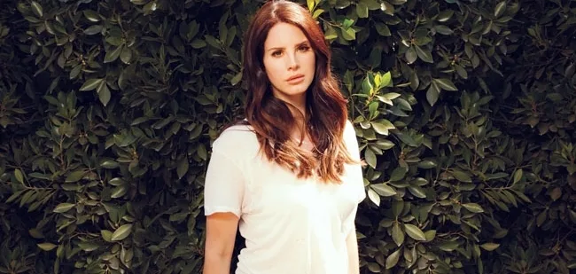 Η Lana Del Rey ποζάρει μπροστά από έναν τοίχο με πυκνή πράσινη φυλλωσιά.