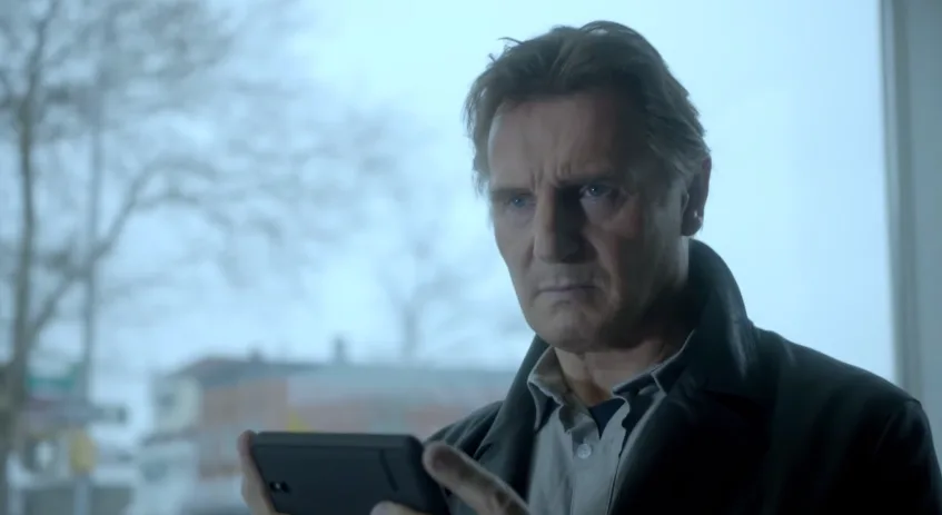 Ο Liam Neeson κοιτάζει με σοβαρό ύφος το κινητό του τηλέφωνο.