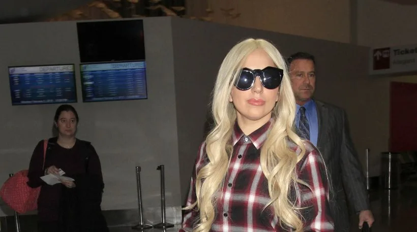 Η Lady Gaga με ξανθά μαλλιά, γυαλιά ηλίου και καρό πουκάμισο στο αεροδρόμιο.