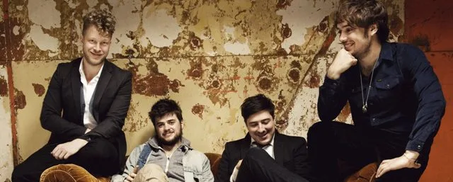 Το συγκρότημα Mumford and Sons ποζάρει μπροστά από έναν φθαρμένο τοίχο.