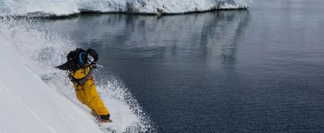 Snowboarder με κίτρινη στολή κάνει κατάβαση σε χιονισμένη πλαγιά δίπλα στη θάλασσα στην Ανταρκτική.
