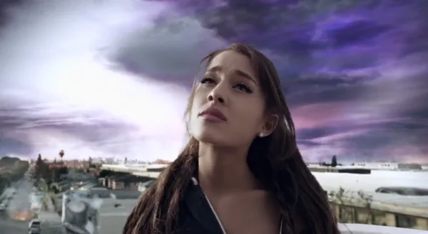 Η Ariana Grande κοιτάζει ψηλά με φόντο έναν μωβ ουρανό και μια πόλη.