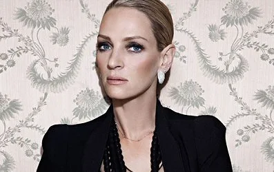Η Uma Thurman με μαύρο σακάκι και κολιέ μπροστά από φλοράλ ταπετσαρία.