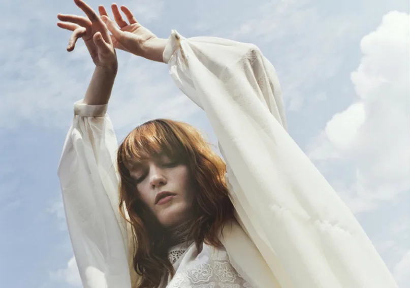 Η Florence Welch με λευκό φόρεμα και υψωμένα χέρια στον ουρανό.