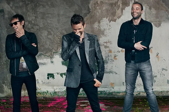 Το συγκρότημα Muse ποζάρει μπροστά από έναν φθαρμένο τοίχο.