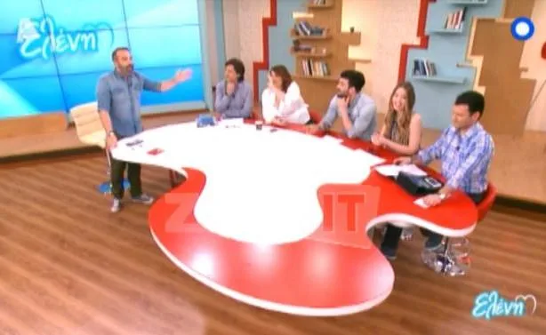 Συνεργάτες της Ελένης Μενεγάκη σε τηλεοπτικό πλατό γύρω από ένα κόκκινο και λευκό τραπέζι.