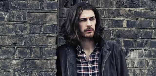 Ο Hozier ποζάρει μπροστά από έναν τοίχο με τούβλα.