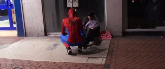 Άνδρας ντυμένος Spiderman σκύβει δίπλα σε άστεγο στο δρόμο τη νύχτα.