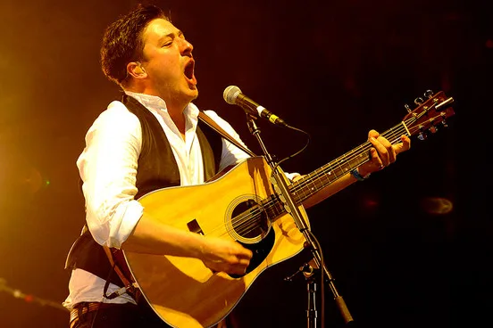 Ο Marcus Mumford των Mumford and Sons τραγουδάει παίζοντας ακουστική κιθάρα στη σκηνή.