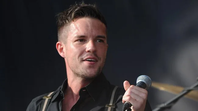 Ο Brandon Flowers τραγουδάει σε σκηνή κρατώντας μικρόφωνο.