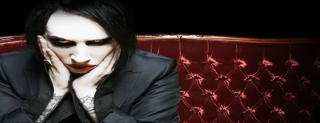 Ο Marilyn Manson με μαύρο κοστούμι και έντονο μακιγιάζ κάθεται σε κόκκινο καναπέ.