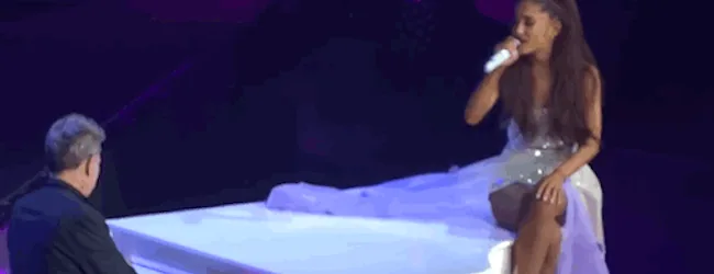 Η Ariana Grande τραγουδάει καθισμένη πάνω σε ένα λευκό πιάνο.