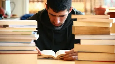 Νεαρός φοιτητής διαβάζει ανάμεσα σε στοίβες βιβλίων.