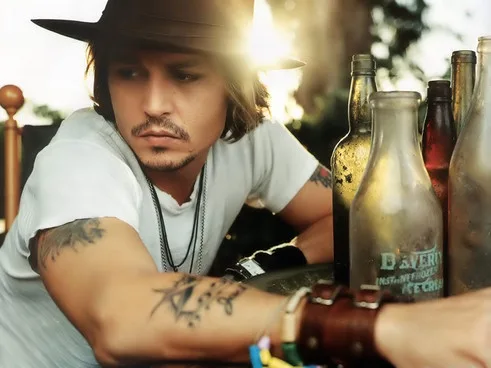 Ο Johnny Depp με καπέλο και τατουάζ κάθεται δίπλα σε γυάλινα μπουκάλια.