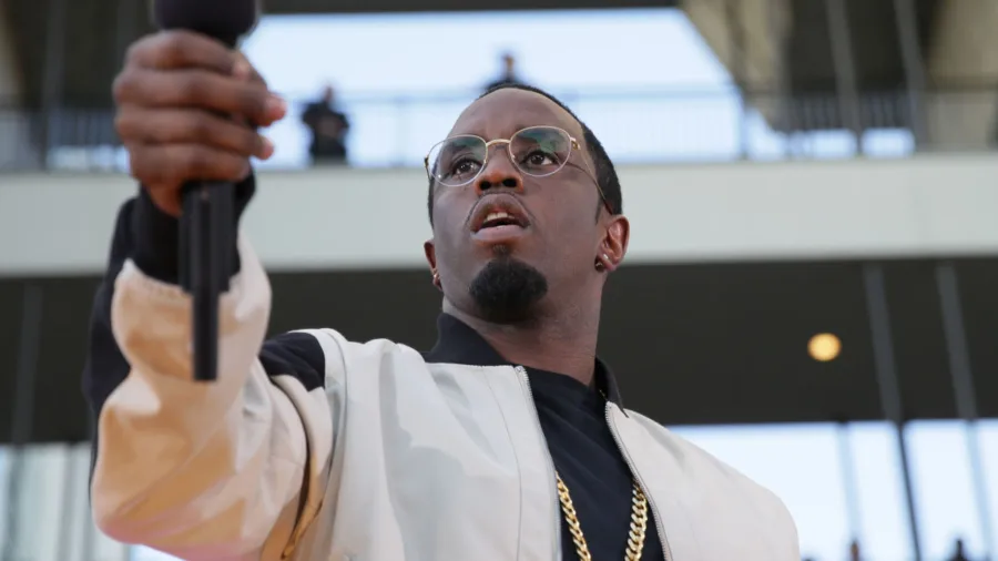 Ο Puff Daddy με γυαλιά και λευκό τζάκετ κρατάει ένα μικρόφωνο.