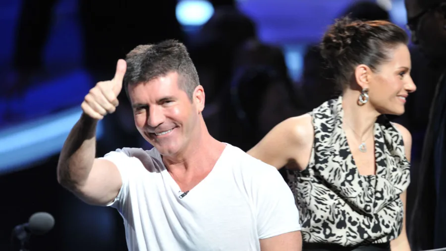 Ο Simon Cowell χαμογελάει και κάνει το σήμα της νίκης με τον αντίχειρα.