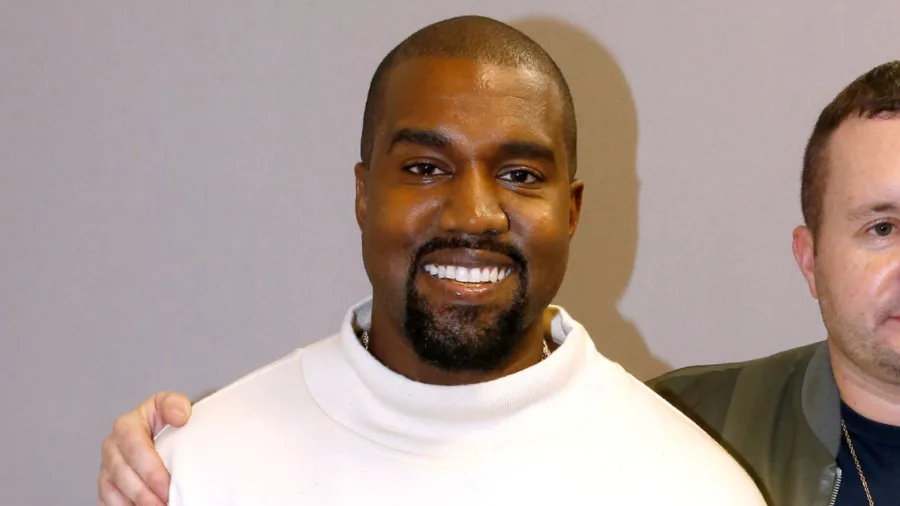 Ο Kanye West χαμογελάει φορώντας λευκό ζιβάγκο.