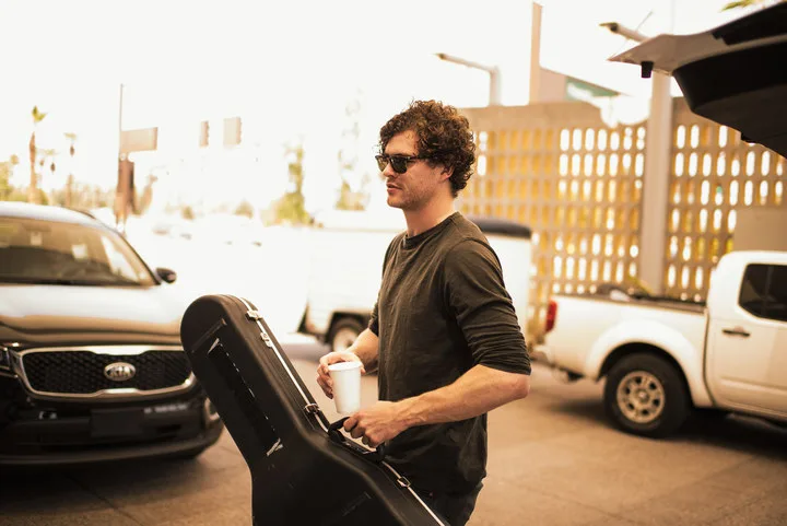 Ο Vance Joy κρατώντας μια θήκη κιθάρας και έναν καφέ δίπλα σε αυτοκίνητα.