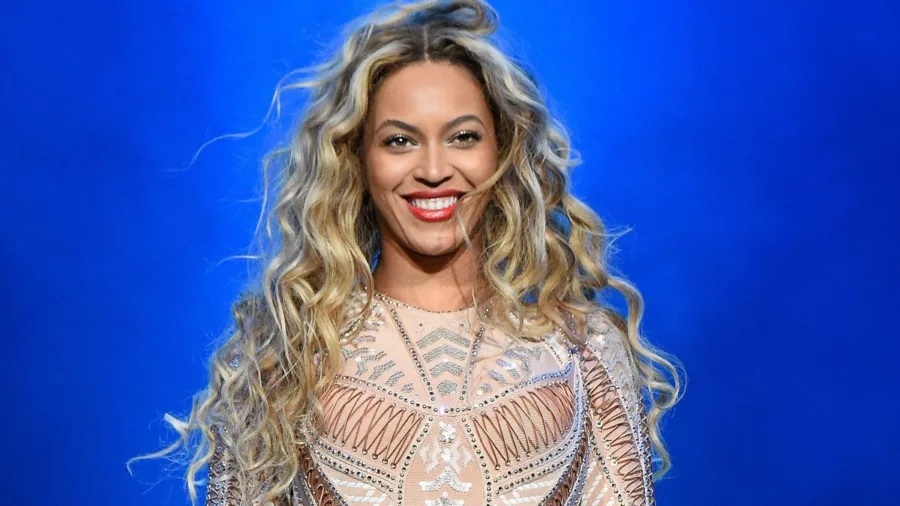 Η Beyoncé χαμογελάει με ξανθά κυματιστά μαλλιά σε μπλε φόντο.
